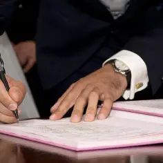 Mężczyzna w garniturze podpisuje dokumenty, być może akt małżeństwa, zgodnie z art. 231 kk. Na ręce ma zegarek.