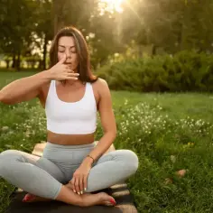 Kobieta w pozycji lotosu ćwiczy oddech, by pokonać zmęczenie psychiczne. Słońce przebija się przez drzewa.