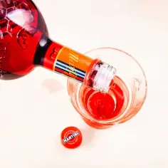 Czerwony Martini Rosso leje się z butelki do szklanki. Na blacie leży czerwona zakrętka z logo Martini.