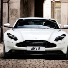 Biały Aston Martin DB11 z tablicą AMV 8 stoi na brukowanej uliczce, otoczony kamiennymi budynkami.