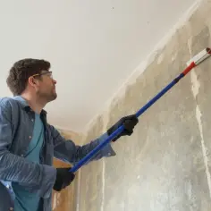 Mężczyzna w okularach ochronnych i rękawiczkach nakłada grunt wałkiem na ścianę. Czy trzeba gruntować przed szpachlowaniem? Tak, to ważny etap przygotowania powierzchni.