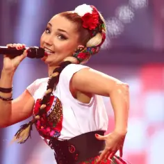 Cleo na Eurowizji w ludowym stroju, z warkoczem i kwiatami we włosach, śpiewa do mikrofonu.