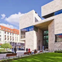 Nowoczesny budynek Teatru Capitol z kamienną fasadą i dużymi oknami. W pobliżu widać starszą kamienicę i ludzi odpoczywających na ławkach.