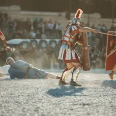 Gladiatorzy w zbrojach toczą zaciętą walkę, jeden z nich leży na ziemi, podczas gdy inni przygotowują się do ataku.