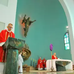 Nabożeństwo w kościele. Ksiądz w czerwonej szacie przy pulpicie, obok ministrant. W tle figura Jezusa i obraz. To może być tekst o męce pańskiej.