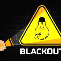 Ręka trzyma latarkę, oświetlając znak ostrzegawczy z pękniętą żarówką. Pod spodem napis "BLACKOUT".
