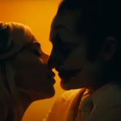 Joker i Harley w namiętnym pocałunku, ich twarze zlewają się w mroku, otoczeni pomarańczowym blaskiem.