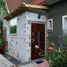 Trwa rozbudowa domu, widać nowe ściany z białych pustaków, drzwi i okno. Wokół kwitną kwiaty.