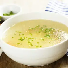 Aromaticzny rosół z perliczki, ozdobiony natką pietruszki, podany z kromką chleba i łyżeczkami.