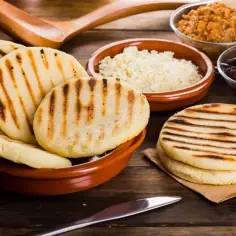 Grillowane arepas w glinianej misce, obok miseczki z białym serem, czarną fasolą i czymś pomarańczowym.