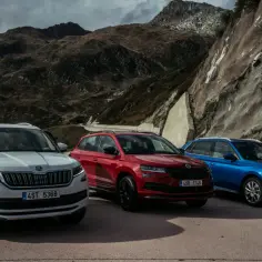 Trzy skoda suv w różnych kolorach: biały, czerwony i niebieski, zaparkowane na tle górskiego krajobrazu.