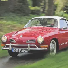 Czerwony Karmann Ghia z białym dachem pędzi po mokrej drodze wśród zieleni.