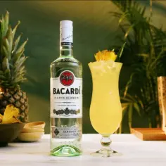 Butelka białego rumu Bacardi obok koktajlu Piña Colada, z ananasami i limonkami w tle.