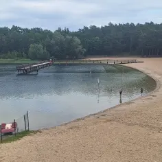 Drewniany pomost nad jeziorem Kamionki, piaszczysta plaża, łódki i dwie osoby kąpiące się w wodzie.