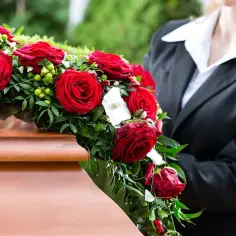 Wieniec z czerwonych róż na drewnianej trumnie, obok osoba w czarnym garniturze.