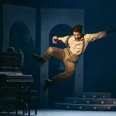 Aktor w podskoku na scenie, w tle pianino i schody. Scena z musicalu "Deszczowa piosenka".