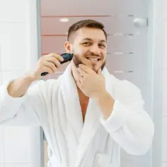 Uśmiechnięty mężczyzna w białym szlafroku goli brodę trymerem w łazience.