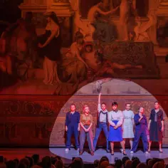 Grupa kobiet na scenie Teatru Narodowego w Warszawie. W tle wielki obraz, z przodu czerwony samochód.