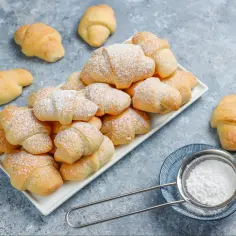 Stos szybkich rogalików kruchych, posypanych cukrem pudrem, obok sitka z cukrem.