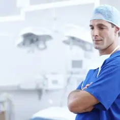 Mężczyzna w niebieskim fartuchu i czepku chirurgicznym stoi ze skrzyżowanymi rękami w jasnej sali operacyjnej.