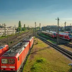 Czerwone lokomotywy i wagony stoją na torach. W tle budynki i drzewa. To miejsce, gdzie przygotowuje się kolej dużych prędkości do podróży.