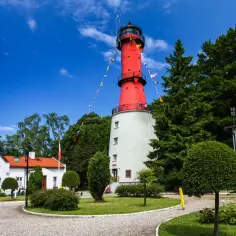 Czerwona latarnia morska, "gwiazda północy" na Jastrzębiej Górze, otoczona zielenią i budynkami.