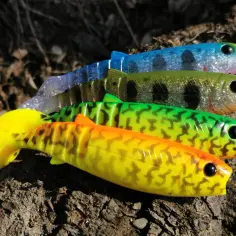 Cztery gumowe przynęty wędkarskie Savage Gear Cannibal w różnych kolorach leżą na korze drzewa.