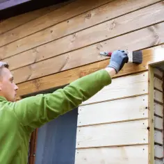 Mężczyzna w zielonej bluzie maluje pędzlem drewnianą elewację domu z deski elewacyjnej, nadając jej nowy wygląd.