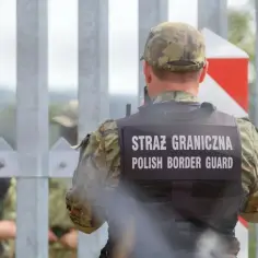 Strażnik graniczny w kamizelce z napisem "STRAŻ GRANICZNA POLISH BORDER GUARD" stoi przy płocie. W tle widać innych funkcjonariuszy i znak graniczny. To obraz polskiej ochrony granic z Rosją.