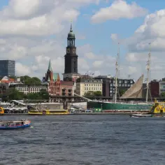 Widok na port w Hamburgu, gdzie statki i łodzie pływają po wodzie, a w tle widać miasto z wieżą kościelną. Czy Hamburg leży nad morzem? Tak, to miasto portowe!