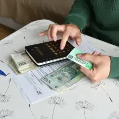Dłonie liczą pieniądze i trzymają legitymację ZUS na tle dokumentów i kalkulatora.