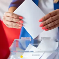 Kobieta z pomarańczowym lakierem na paznokciach wrzuca kartę do urny wyborczej. W tle polskie flagi, symbolizujące głosowanie za granicą.