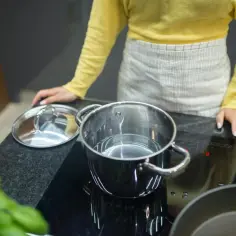 Kobieta w fartuchu i żółtej bluzce przygotowuje wodę w garnku na płycie indukcyjnej. Czerwony wskaźnik pokazuje, co oznacza "L" na indukcji - niski poziom mocy.