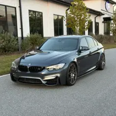 Szary sedan BMW M3 2025 z przyciemnianymi szybami i felgami stoi przed nowoczesnym budynkiem.
