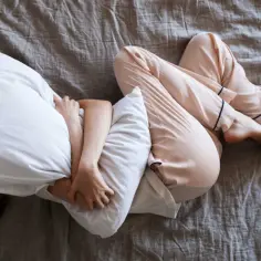 Osoba w piżamie skulona na łóżku, przytulająca poduszkę. Ciało wygięte w łuk, ręce obejmujące głowę, symbolizujące wyuczoną bezradność.