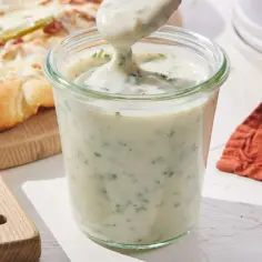 Gęsty, kremowy biały sos do pizzy z ziołami w słoiku, obok gotowej pizzy. Idealny dodatek do każdej pizzy.