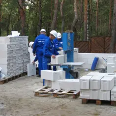 Na budowie pracownicy w niebieskich kombinezonach i białych kaskach przygotowują pustaki. Widać wiele palet z pustakami, które czekają na ułożenie.