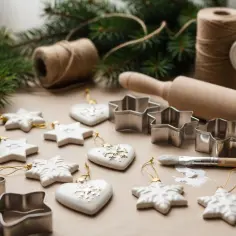 Białe, porcelanowe ozdoby choinkowe w kształcie gwiazdek i serc, metalowe foremki, pędzel i wałek do ciasta sugerują tworzenie świątecznych dekoracji z masy porcelanowej.