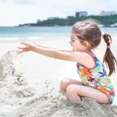 Dziewczynka w kolorowym stroju kąpielowym z okularami na nosie rzuca piasek, ciesząc się zabawami na plaży.