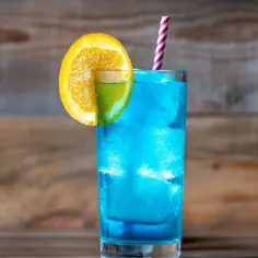 Orzeźwiający blue lagoon drink przepis. Błękitny koktajl z lodem, plasterkiem pomarańczy i limonki, ozdobiony słomką.