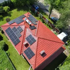 Dach z panelami fotowoltaicznymi, które stanowią dodatek energetyczny dla domu. Widać też samochody i zieleń.