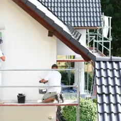 Dwóch malarzy na rusztowaniu maluje elewację domu. Sprawdź, jaka jest malowanie podbitki cena i odśwież swój dom.