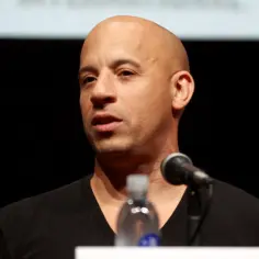 Vin Diesel, znany z imponującego wzrostu, w czarnej koszulce przemawia do mikrofonu.