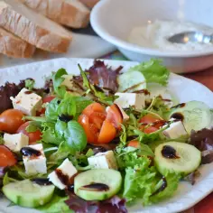 Świeża sałatka z pomidorami, ogórkiem i serem feta, polana aromatycznym sosem truflowym. Obok kromki chleba i miseczka dipu.