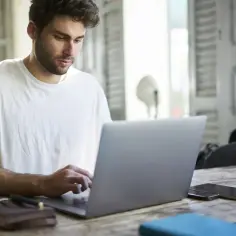 Mężczyzna w białej koszulce pracuje na laptopie, szukając pracy. Na stole leży notatnik i długopis.