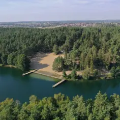 Malownicza borówno plaża otoczona lasem, z drewnianymi pomostami wchodzącymi do ciemnej wody. W oddali widać zabudowania.