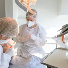 Dentystka w masce i fartuchu wykonuje zabieg kiretażu u pacjentki. W tle widać sprzęt stomatologiczny.