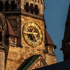 Złoty zegar na wieży kościoła pamięci cesarza Wilhelma, oświetlony słońcem, góruje nad kamienną architekturą.