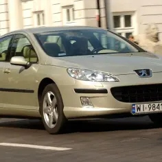 Złoty Peugeot 407 w ruchu miejskim. Kierowca w środku, budynki w tle. Wiele osób szuka opinii o tym modelu.