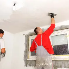 Dwóch mężczyzn w kombinezonach remontowych tynkuje sufit i ściany w pomieszczeniu z oknem.